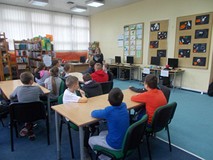 Dzieci siedzą przy stołach i słuchają opowiadania czytanego przez bibliotekarkę