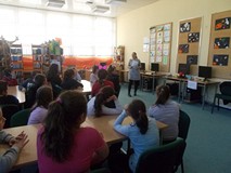 Uczniowie siedzą przy stołach i słuchają bibliotekarki czytającej książki