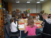 Dzieci siedzą przy stole i wykonują prace plastyczną, a bibliotekarz wpsomaga ich pracę