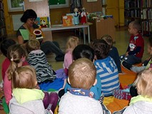 Dzieci siedzą na chuście integracyjnej, bibliotekarka pokazuje książeczki z zagadkami