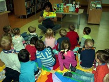 Dzieci siedzą na chuście integracyjnej, bibliotekarka pokazuje książeczki z zagadkami
