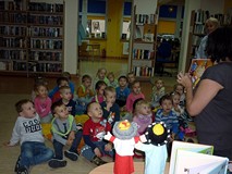 Dzieci siedzą na chuście integracyjnej, bibliotekarka pokazuje ilustracje do znanych baśni