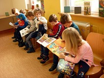 Dzieci siedzą na krzesełkach i przeglądają książki z wystawki BAJKOWE ŚWIĘTA