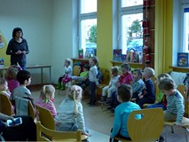 Dzieci siedzą na krzesełkach, bibliotekarka wita się z grupą