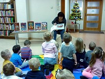 Dzieci siedzą na chuście animacyjnej i słuchają czytanej przez bibliotekarkę książeczki ŚWIĘTA PANA 