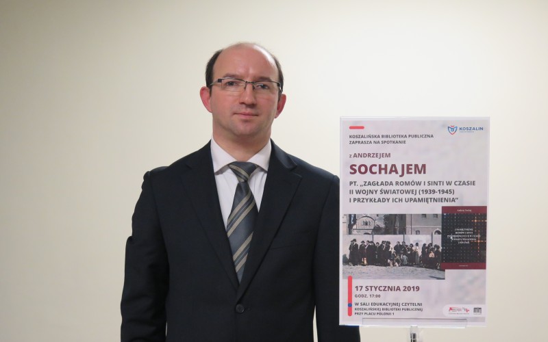 Andrzej Sochaj obok tabliczki z platatem promującym wykład na temat zagłady Romów i Sinti