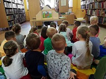 Dzieci siedzą na chuście i słuchają czytającej  bibliotekarki.