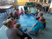 Dzieci siedza na dywanie i słuchają czytanej przez bibliotekarkę książki