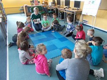 Dzieci siedza na dywanie i słuchają czytanej przez bibliotekarkę książki