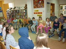Dzieci siedzą na krzesełkach i słuchają czytającej  bibliotekarki.