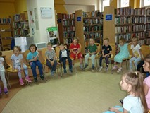 Dzieci siedzą na krzesełkach i słuchają czytającej  bibliotekarki.