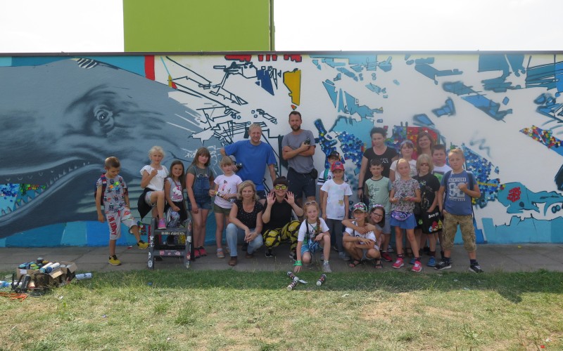 Pamiątkowe zdjęcie. Na tle muralu z Moby Dickiem stoją dzieci, które pomagały przy jego...