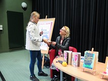 Uczestniczka spotkania odbiera ksiazkę z autografem od Joanny Jagiełło. Dziewczynka stoi, autorka si