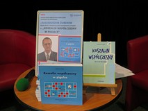 Plakat promujący spotkanie oraz dwie ksiązki autora są wyeksponowane na okrągłym stoliku.