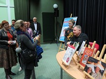 Robert Makłowicz siedzi przy stoliku i rozdaje autografy. Otaczają go uczestnicy spotkania.