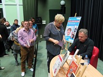 Robert Makłowicz siedzi przy stoliku i rozdaje autografy. Otaczają go uczestnicy spotkania.