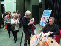 Robert Makłowicz siedzi przy stoliku i rozdaje autografy. Otaczają go uczestnicy spotkania.