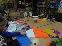 Dzieci siedzą przy stole i wykonują prace plastyczną, podczas gdy bibliotekarka czyta dzieciom opowi