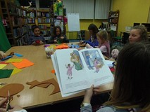 Dzieci siedzą przy stole i wykonują prace plastyczną, podczas gdy bibliotekarka czyta dzieciom opowi