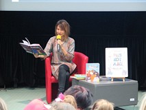 Agata Widzowska siedzi na czerwonym fotelu przy szarym stoliku i czyta fragment swojej książki z ser