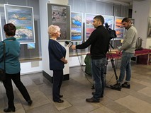 Ewa Maliszewska udziela wywiadu, w galerii na tle swoich obrazów, z lewej strony widać oglądającego 