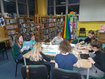 Dzieci siedzą przy stole i wykonują prace plastyczną, podczas gdy bibliotekarka czyta opowieść