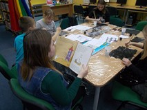 Dzieci siedzą przy stole i wykonują prace plastyczną, podczas gdy bibliotekarka czyta opowieść