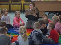 Dzieci siedzą na chuście animacyjnej i słuchają czytnego przez bibliotekarkę opowiadania