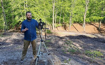 Archeolog Andrzej Kuczkowski stoi w lesie, przed nim na statywie umieszczony jest aparat