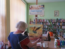 Na tle witryny okiennej i regałów z książkami stoi tyłem Teresa Turkiewicz obok sztalugi z namalowan
