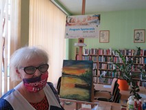 Na tle witryny okiennej i regałów z książkami Teresa Turkiewicz stoi obok sztalugi z namalowanym prz
