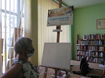 Na tle witryny i regałów z książkami Iwona Sołoducho stoi obok sztalugi z płótnem.