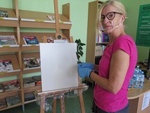 Na tle regałów z prasą stoi Elżbieta Knitter obok sztalugi z płótnem.