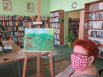 Na tle regałów z książkami stoi sztaluga z obrazem namalowanym przez Agnieszkę Witkowską, obok siedz