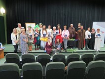 Na zdjęciu:na scenie stoi liczna grupa osób w kolorowych strojach i nakryciach głowy biborących udzi