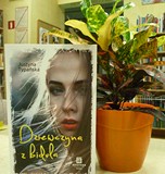 na tle regałów bibliotecznych, obok kwiatka w doniczce stoi książka Dzieczyna z bidula autorstwa Typ