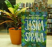 na tle regałów bibliotecznych, obok doniczkowego kwiatka stoi książka Jasna sprawa autorstwa Płatkow