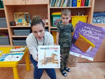 Bibliotekarka pokazuje dzieciom książkę. Z prawej strony stoi chłopiec trzymający w ręce filetowy pl