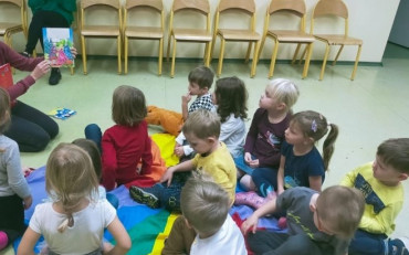 Bibliotekarka pokazuję dzieciom książkę. Dzieci siędzą na kolorowej chuście.