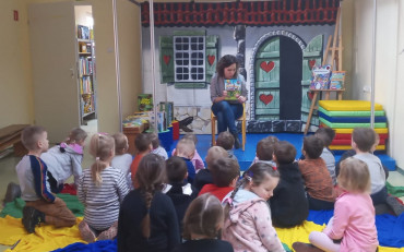 Dzieci siedzą na kolorowej chuście. Pani bibliotekarka pokazuje książkę o dinozaurach.