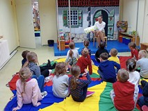 Dzieci siedzą na kolorowej chuście i słuchają książki czytanej przez bibliotekarkę.