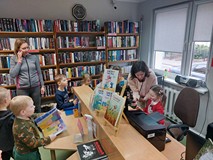 Dziewczynka siedzi za biurkiem i trzyma w ręku książkę. Obok stoi bibliotekarka. W tle dzieci oraz r