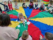 Dzieci siedzą w kole na kolorowej chuście. Jedna dziewczynka zgaduje, co jest pod białymi kartkami