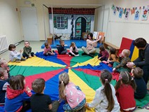 Dzieci podczas zajęć siedzą w kole na kolorowej chuście i słuchają bibliotekarki, czytającej książkę