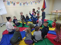 Dzieci siedzą na kolorowej chuście i słuchają książki czytanej przez bibliotekarkę.