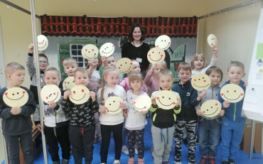 Dzieci stoją na niebieskiej scenie i trzymają w dłoniach żółte papierowe emotikony