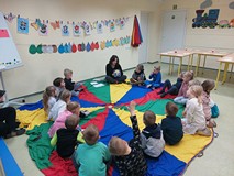 Dzieci podczas zajęć siedzą w kole na kolorowej chuście i słuchają bibliotekarki, czytającej książkę