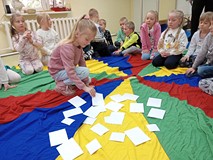 Dzieci siedzą w kole na kolorowej chuście. Jedna dziewczynka zgaduje, co jest pod białymi kartkami