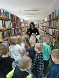 Dzieci stoją między regałami i słuchają bibliotekarki, stojącej przed nimi