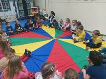 Dzieci podczas zajęć siedzą w kole na kolorowej chuście i słuchają bibliotekarki, czytającej książkę